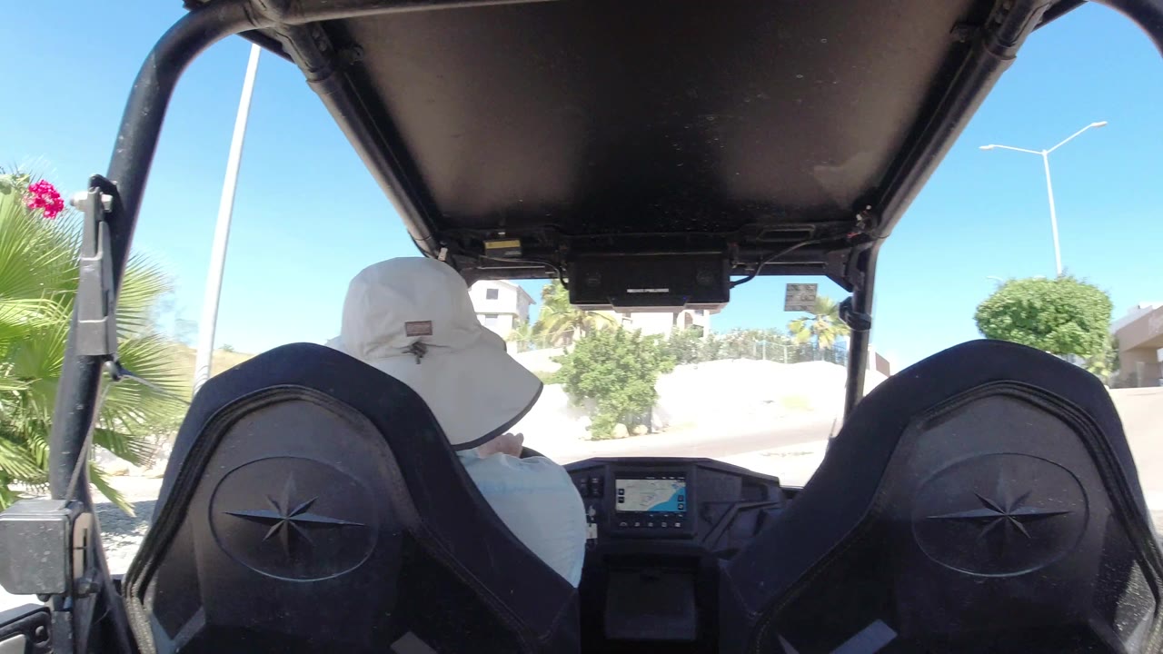The driver remains framed between black Wrangler seats as the bright Los Cabos street, shrubs, a white wall, and a tall streetlight appear beyond the windshield area.