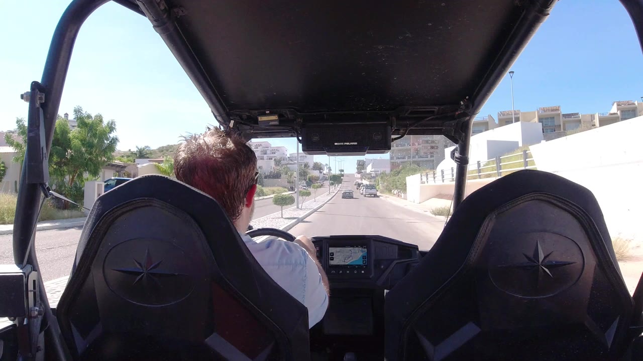 Rear passenger view in Los Cabos, Mexico, looking past black vehicle seats and a driver from behind toward a narrow road lined with white walls and buildings.