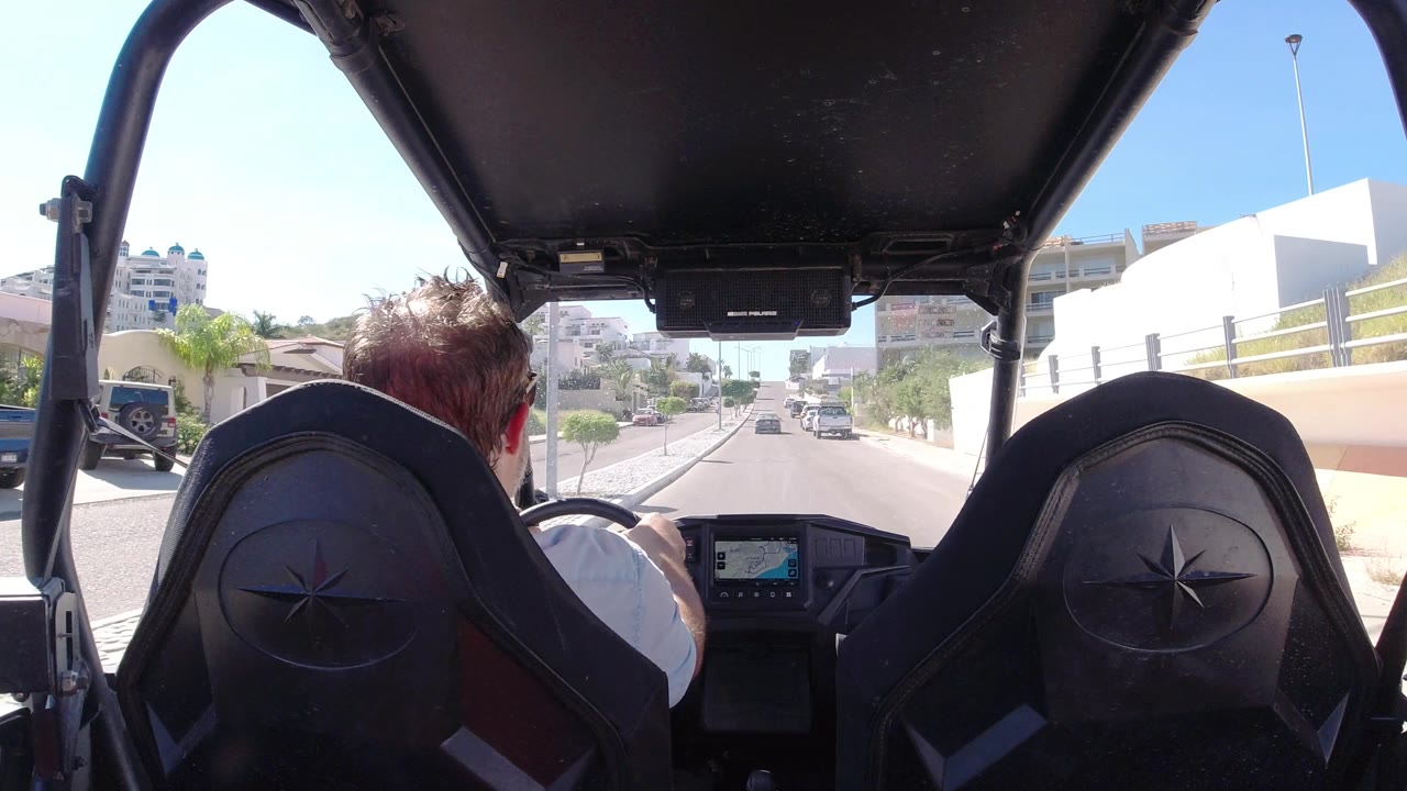 Driver seen from behind as the vehicle approaches parked cars and white buildings.