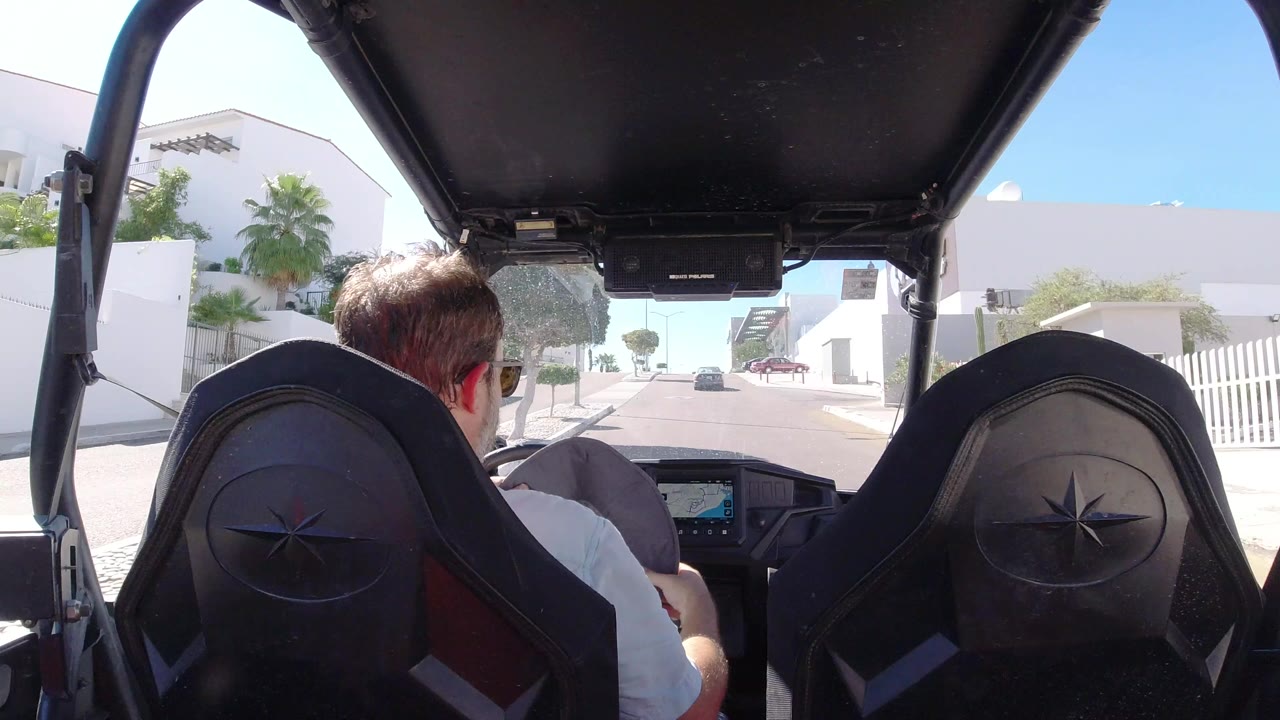 Man driving a golf cart under a black roof on a sunny resort road.