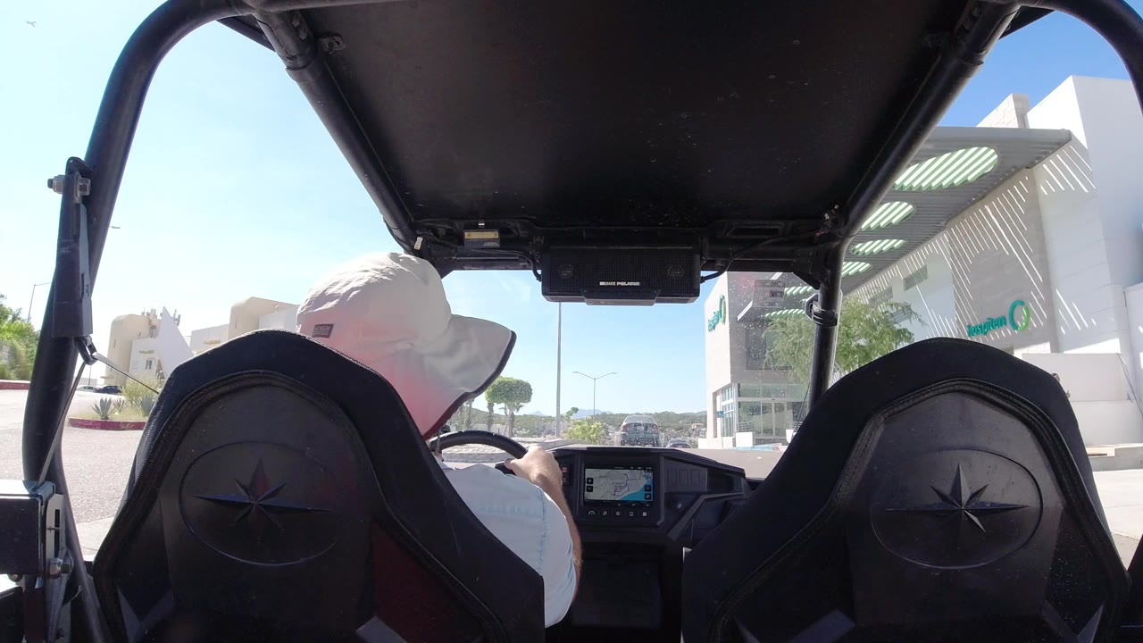 Inside the open vehicle in Los Cabos, the driver remains centered left while a large building, distant cars, and a bright roadway fill the background.