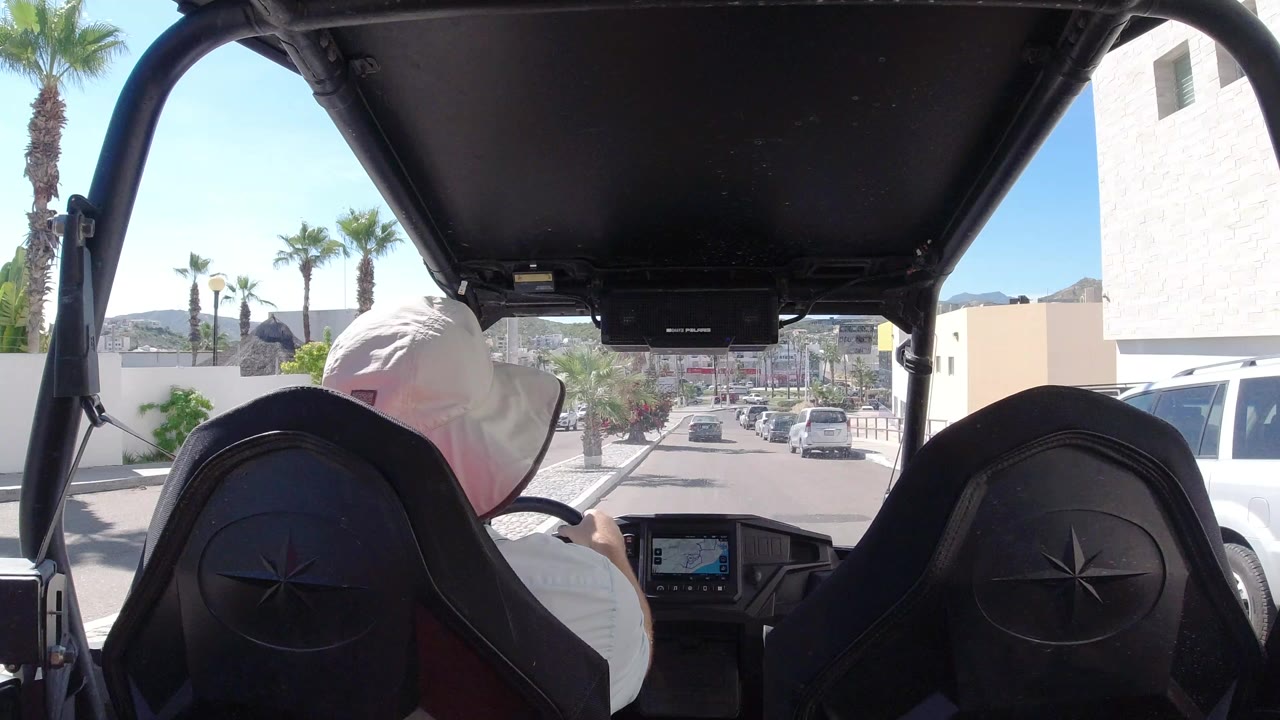Rear passenger view in Los Cabos, Mexico, with a man in a light hat driving an open buggy past parked cars, palms, white buildings, and distant hills.