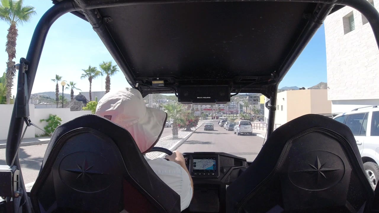 Inside-buggy view in Los Cabos, Mexico, with the driver framed by black seats and roof supports as the street continues between palms, parked vehicles, and pale buildings.