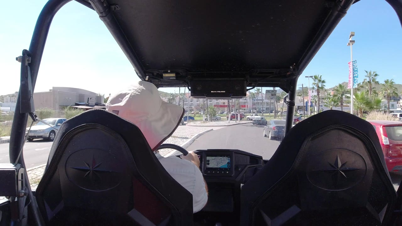 Rear-facing interior view in Los Cabos, Mexico, with the driver leaning slightly left, shaded seats in the foreground, a red parked vehicle at right, palms, and low buildings ahead.