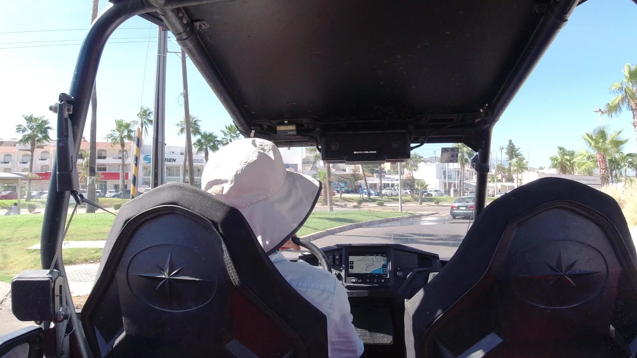 The Los Cabos street remains centered beyond the vehicle dashboard, with the driver turned slightly left, palms along the median, and cars moving through the bright roadway.