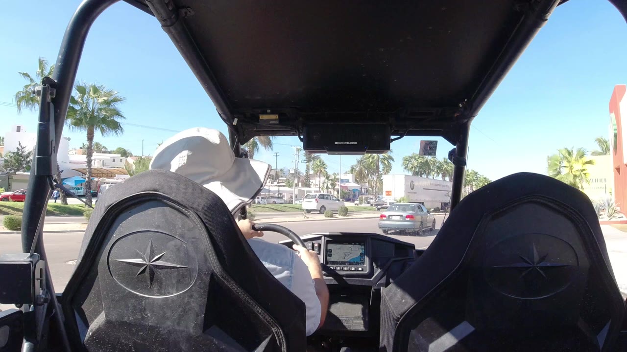 At 0:00, a man in a white hat drives a Wrangler through Los Cabos, Mexico, with black seatbacks framing cars, palms, storefronts, and bright sky ahead.