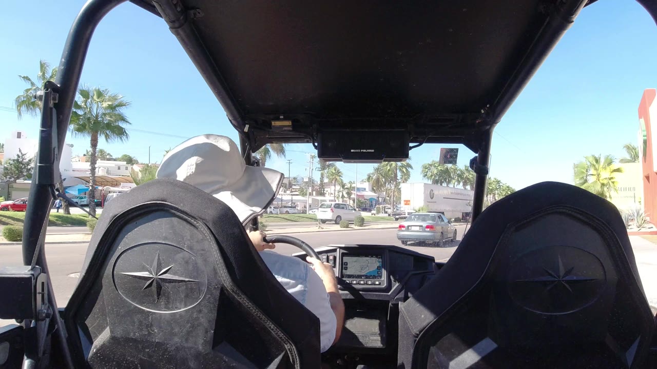 At 0:01, the rear cabin view shows the driver slightly farther along the Los Cabos road, with a gray car ahead, palm trees, low buildings, and strong sunlight outside.