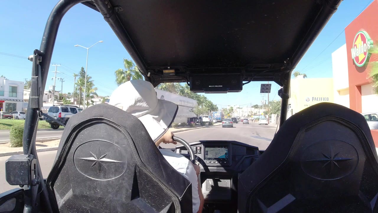 Back-seat view in Los Cabos, Mexico, looking past black off-road seats and a driver in a white hat toward a sunny road with storefronts and traffic.