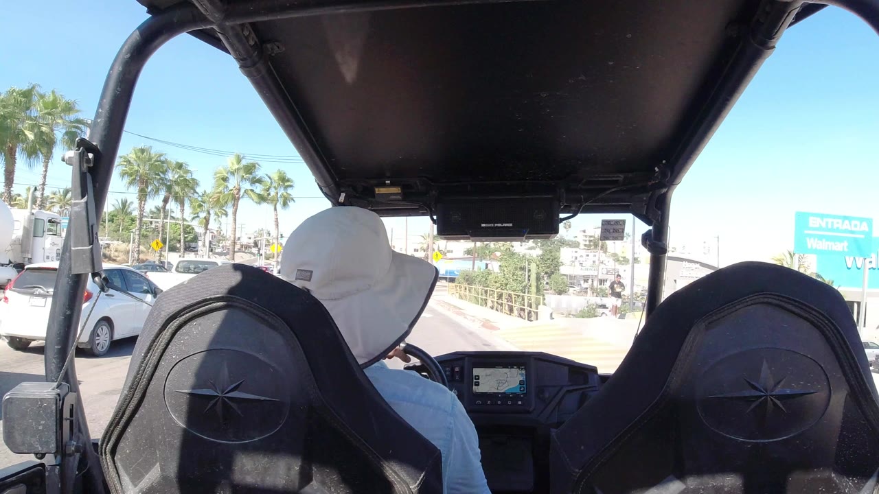 Backseat view from a golf cart approaching a sunny street with palm trees and cars.