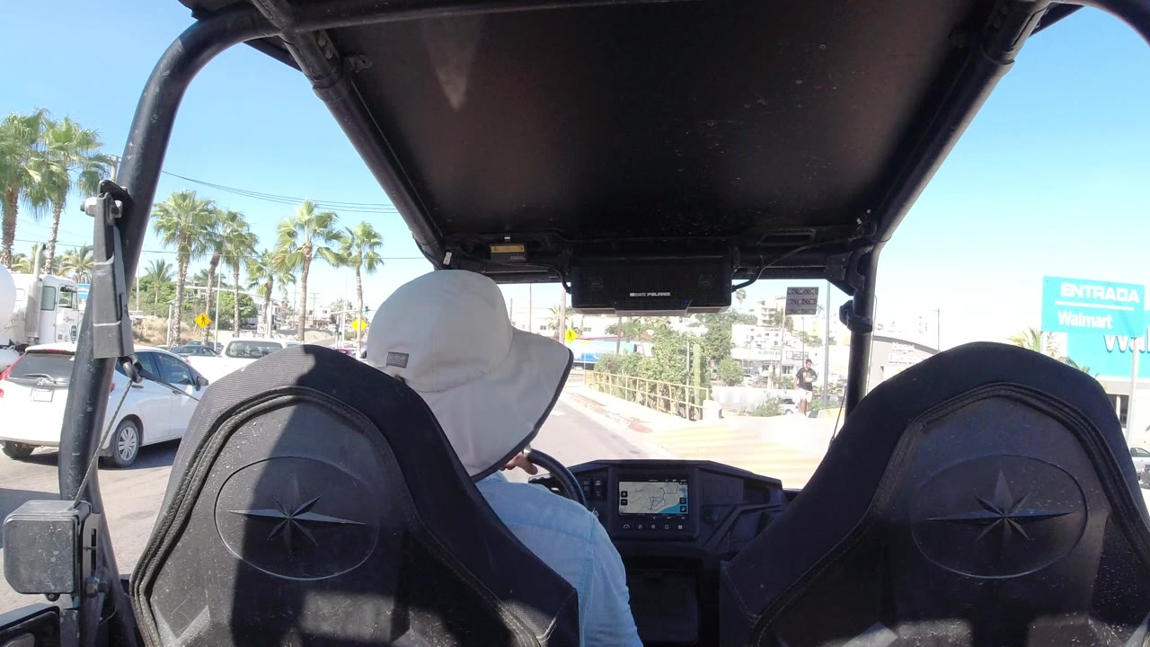 From the rear of the golf cart in Los Cabos, the driver sits ahead under the roof while parked or slow-moving cars, palm trees, a low roadside wall, and Walmart signage fill the sunlit background.