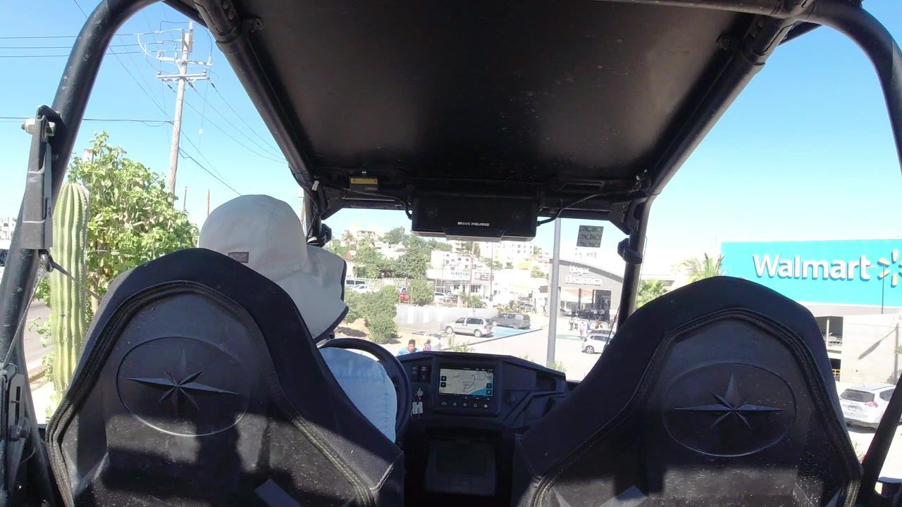 Rear-seat view inside an open vehicle in Los Cabos, Mexico, with black seatbacks, a driver in a white hat, dashboard display, bright sky, parked cars, and Walmart signage ahead.