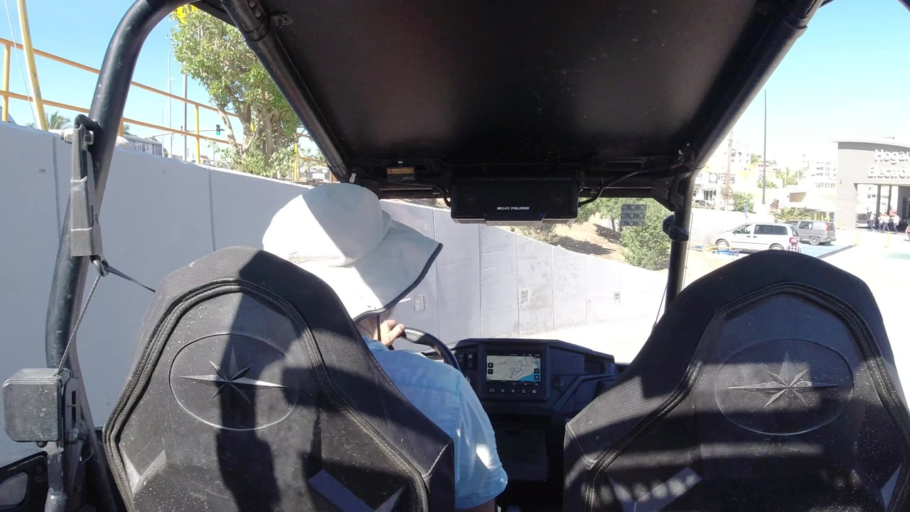 At 0:00 in Los Cabos, Mexico, a hat-wearing driver is seen from behind under a black vehicle roof, with black seatbacks, bright pavement, parked cars, pedestrians, and a retail storefront ahead.