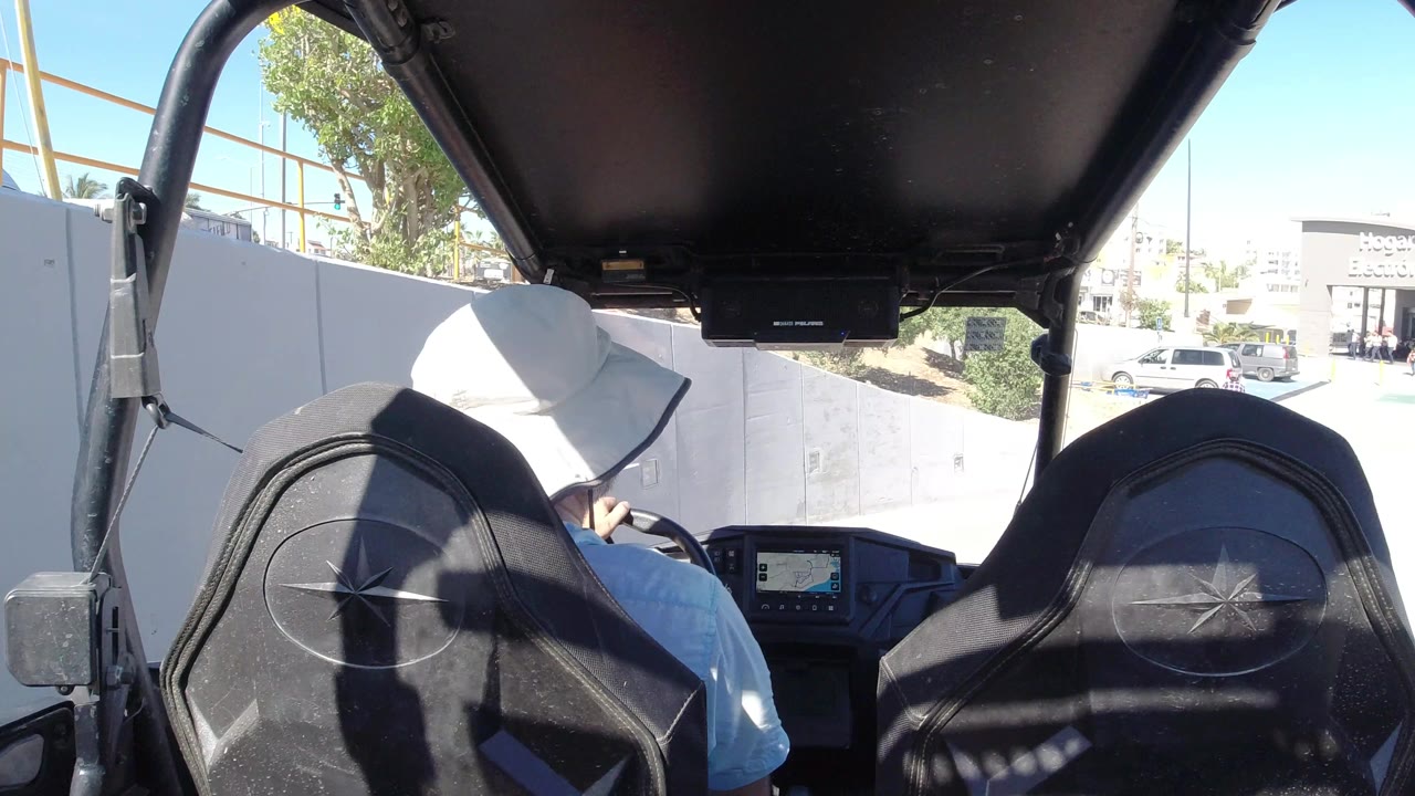 At 0:01 in Los Cabos, Mexico, the driver remains centered beneath the dark roof frame while the vehicle faces a bright parking area with storefronts, cars, and small pedestrians in the distance.
