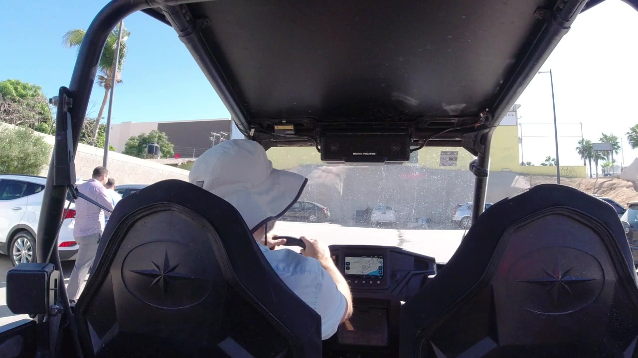 Rear passenger view in Los Cabos shows a man in a white hat at the wheel, black seat backs in the foreground, parked cars, palms, and a pale wall ahead.