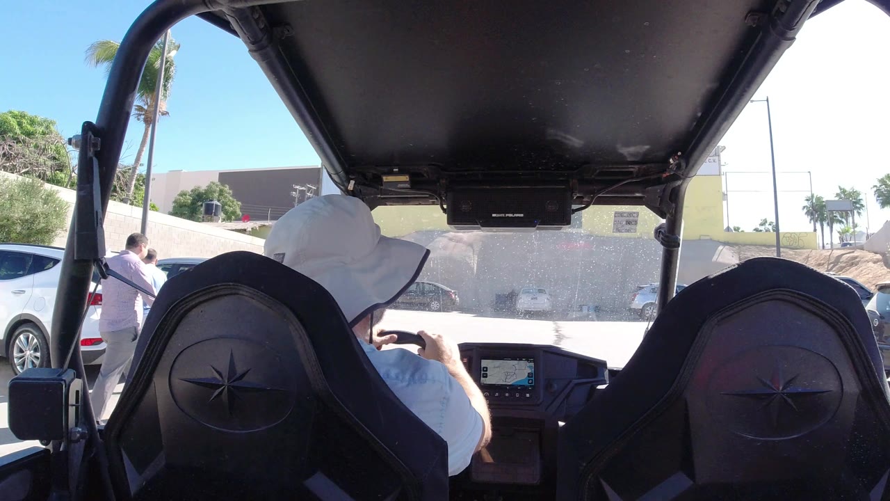 The driver remains centered under the dark canopy as the vehicle faces a bright Los Cabos parking lot with cars along a wall and low buildings beyond.