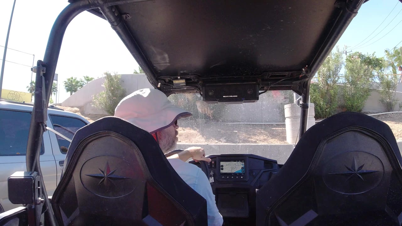 The man remains seated at the wheel in Los Cabos, Mexico, framed by the open vehicle roof and two black seats, with a parked vehicle and sunlit landscaping outside.