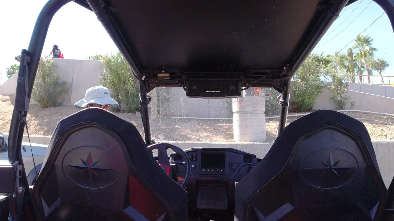 At 0:00 in Los Cabos, Mexico, the rear interior of a vehicle frames two black seats while a man in a white hat stands outside near the left side.