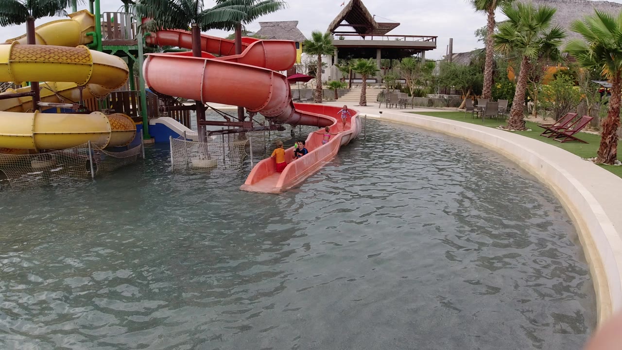 One second in, the riders remain low in the red slide chute while the yellow slide, palm trees, and curved pool border stay prominent in the Los Cabos water park setting.