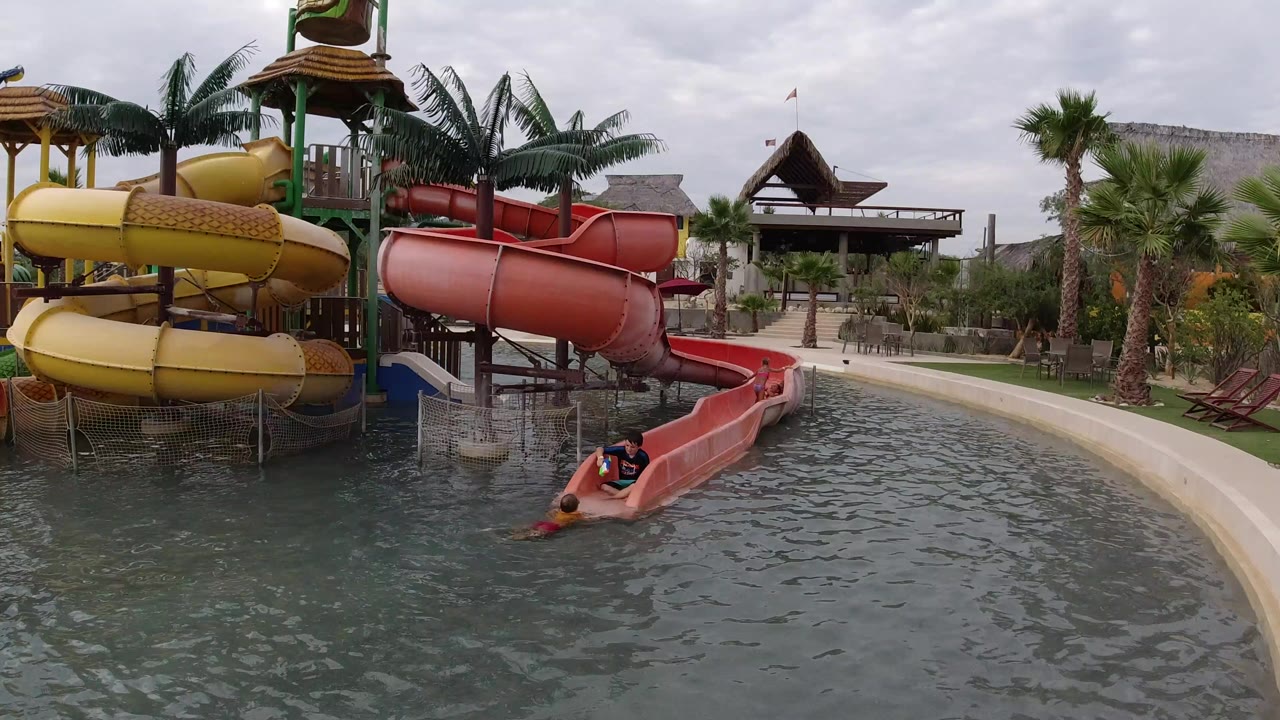 Children are visible near the exit of a red water slide in a Los Cabos, Mexico pool, with yellow spiral slides, palm trees, and cloudy daylight behind them.