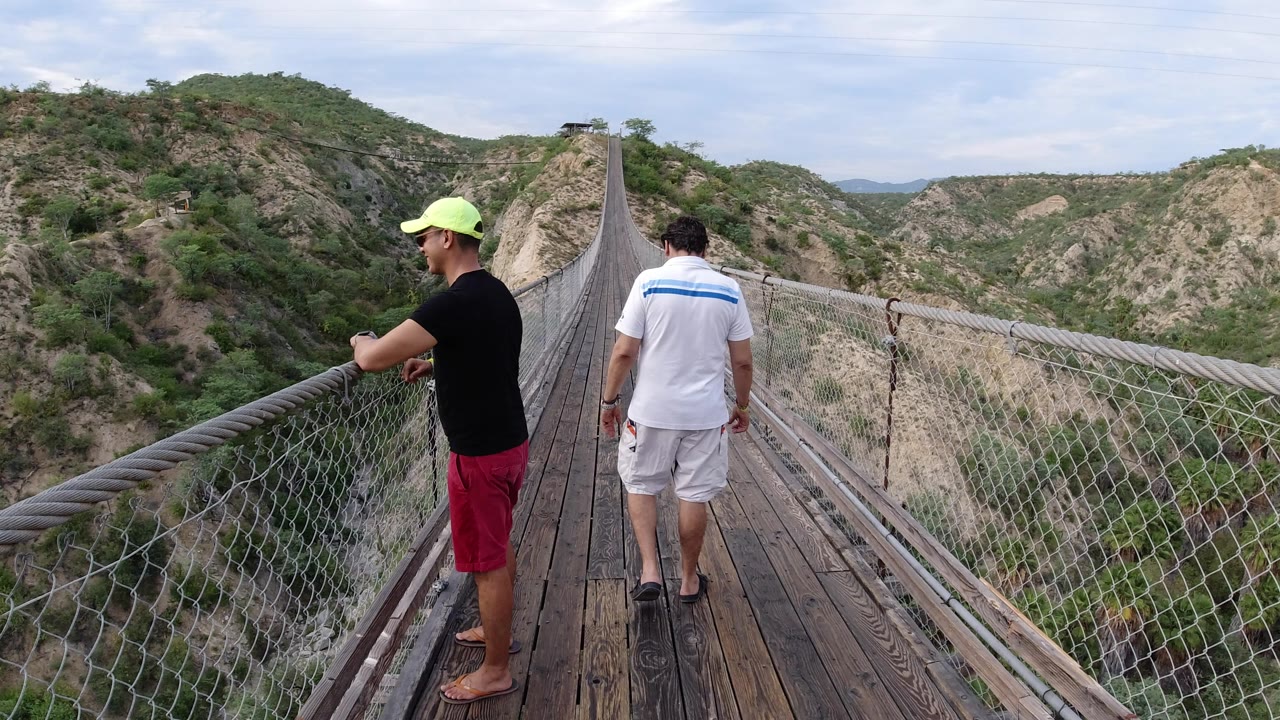 One man in a neon cap stands closer to the camera while another in a white shirt faces down the Los Cabos hanging bridge toward the hills.