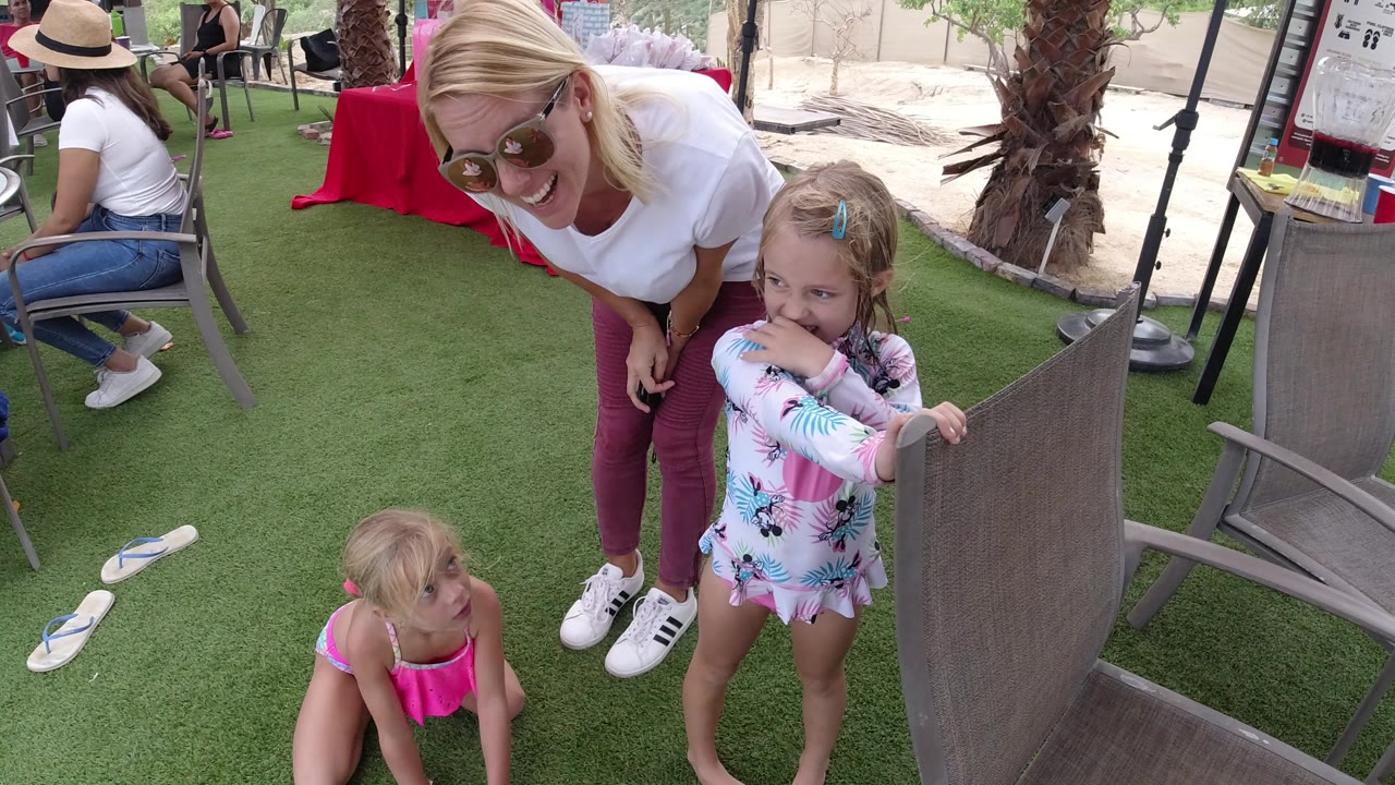 In Los Cabos, Mexico, the woman bends forward smiling as one young girl grips a patio chair and another girl in a pink swimsuit sits on the turf.
