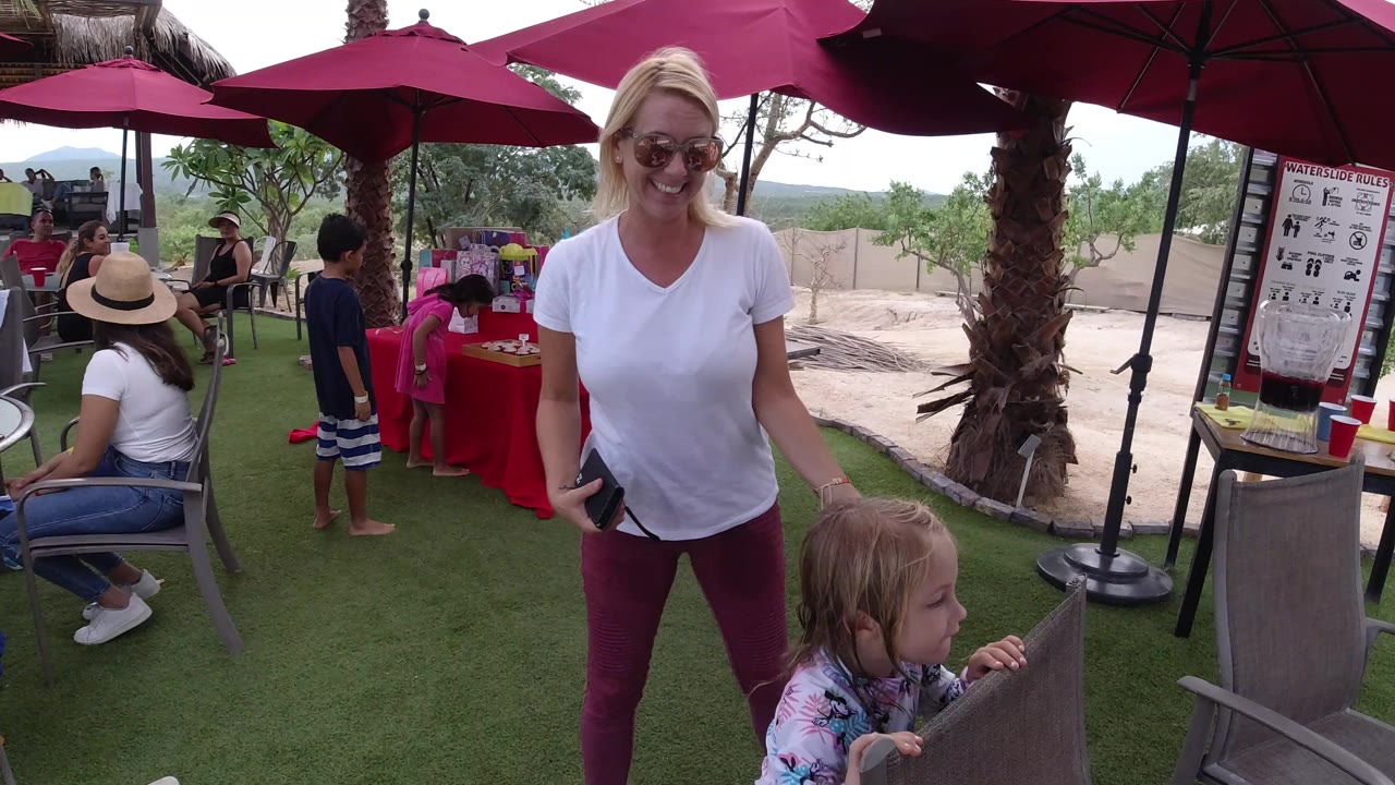 The woman smiles toward the camera as the little girl stands at the edge of the frame, with a red table, seated adults, children, and umbrella shade behind them in Los Cabos.