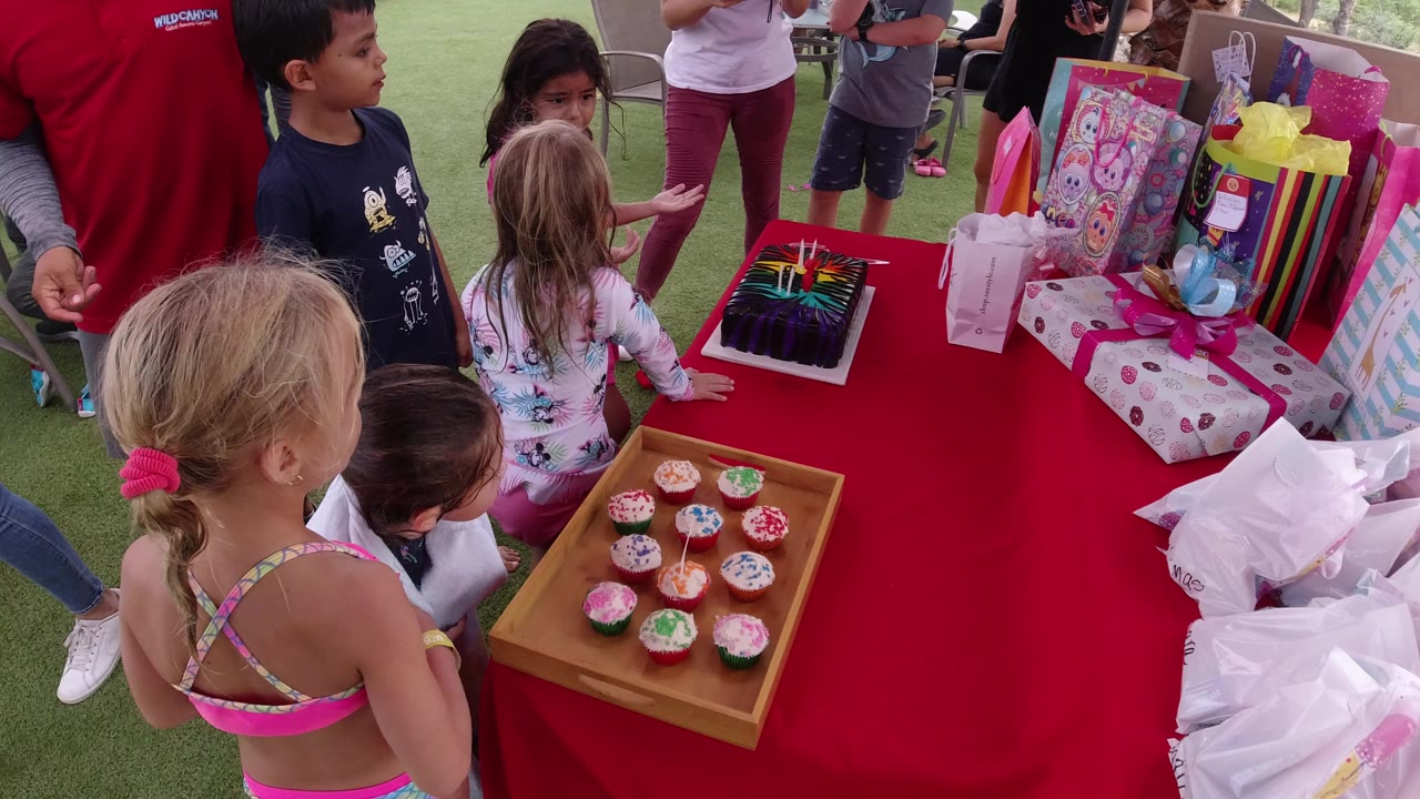 In Los Cabos, Mexico, several children stand around a red party table holding cupcakes, a dark birthday cake, wrapped presents, and gift bags while adults are partly visible nearby.