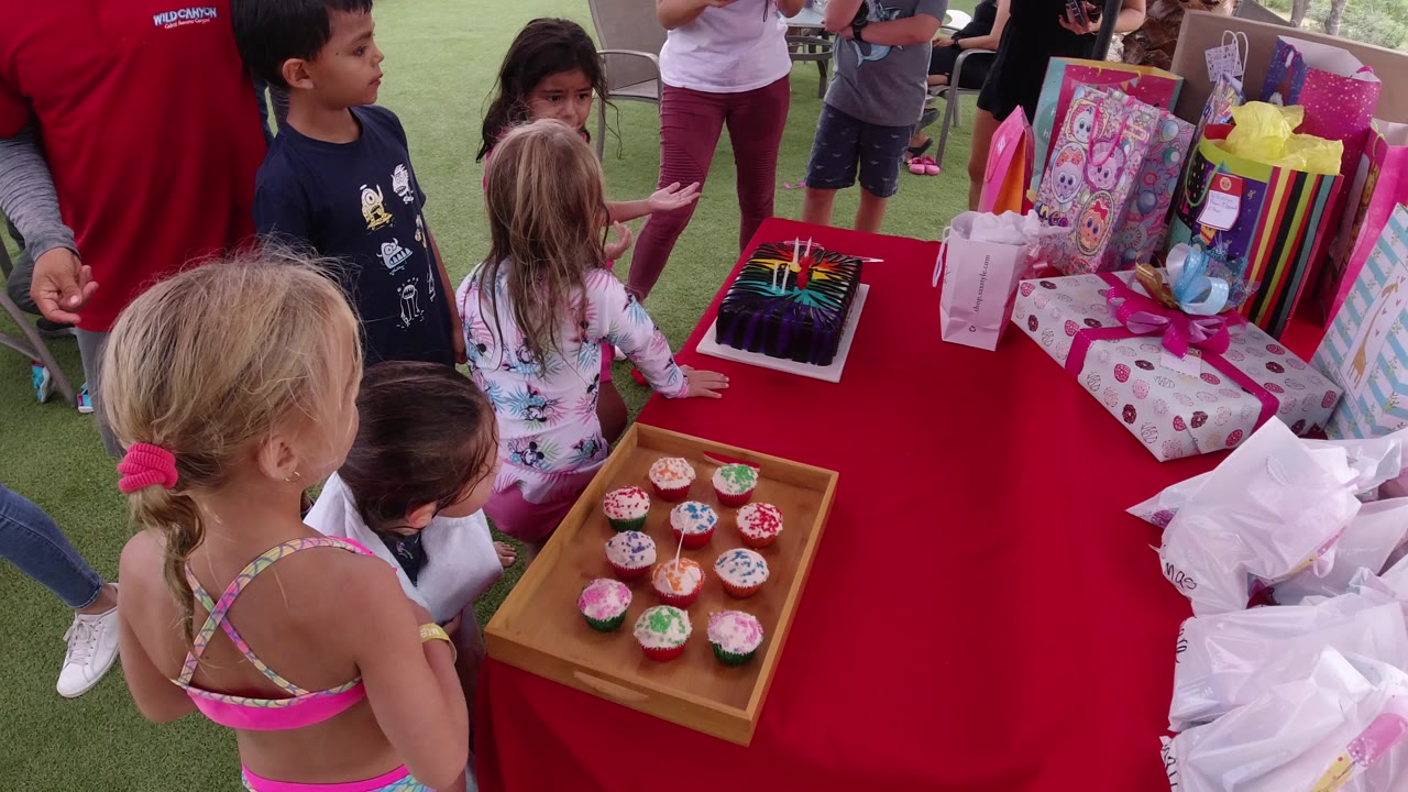 In Los Cabos, Mexico, children remain gathered beside a red table with a tray of frosted cupcakes, a dark cake with colorful decoration, and multiple wrapped gifts.