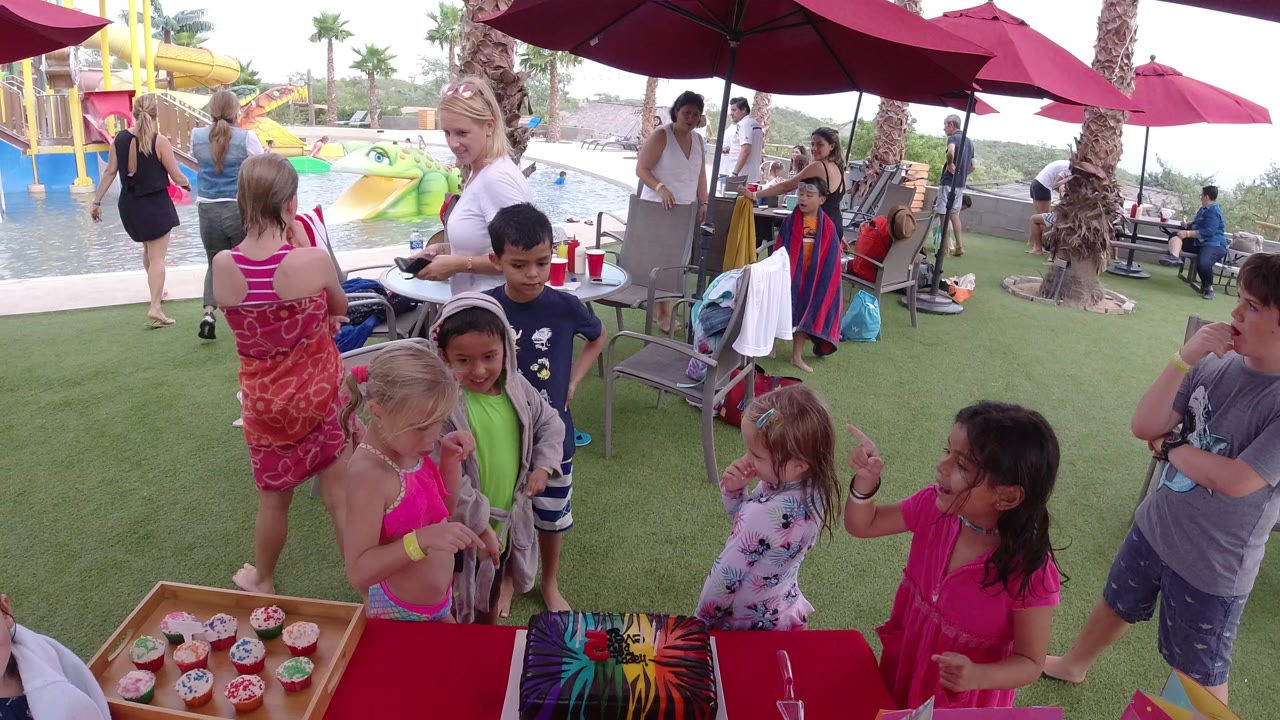 Children and adults gathered around a red party table near a water park