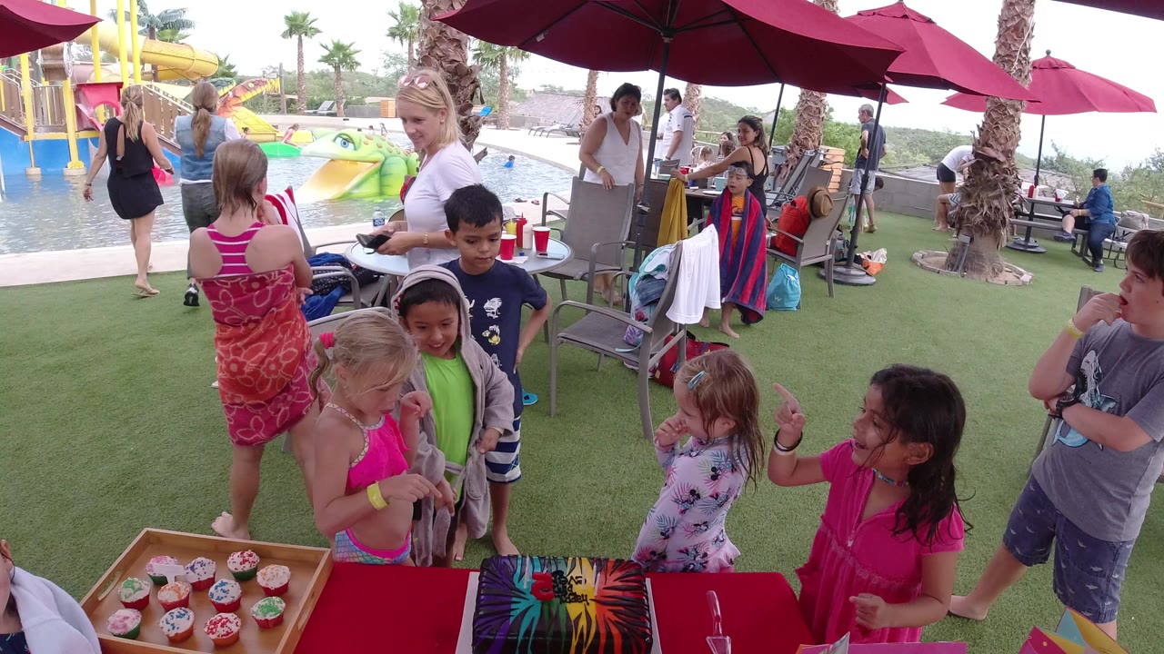 In Los Cabos, several children cluster beside cupcakes and a colorful table covering while adults stand nearby under red umbrellas at a water play area.