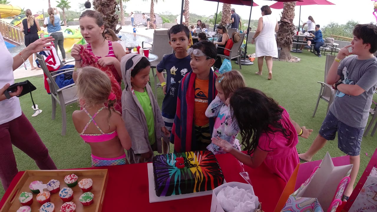A group of children shifts around the cupcake table at the Los Cabos club while an adult in a white shirt stands beside them holding a small item.
