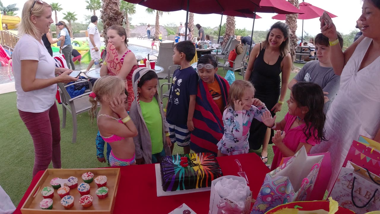 Children in swimwear and towels stand beside a red birthday table in Los Cabos while adults gather around them under red umbrellas near pool-play equipment.