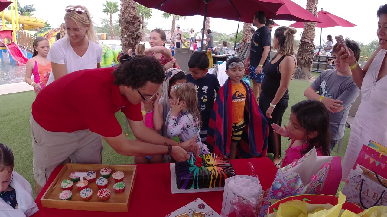 An adult continues reaching over the cake while a girl in pink and other children watch closely at the Los Cabos poolside birthday table.