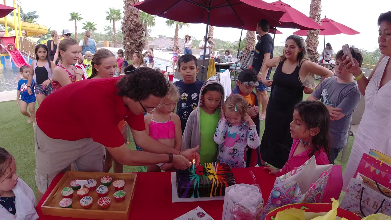 Children gather around a birthday cake as a man leans in with candles.