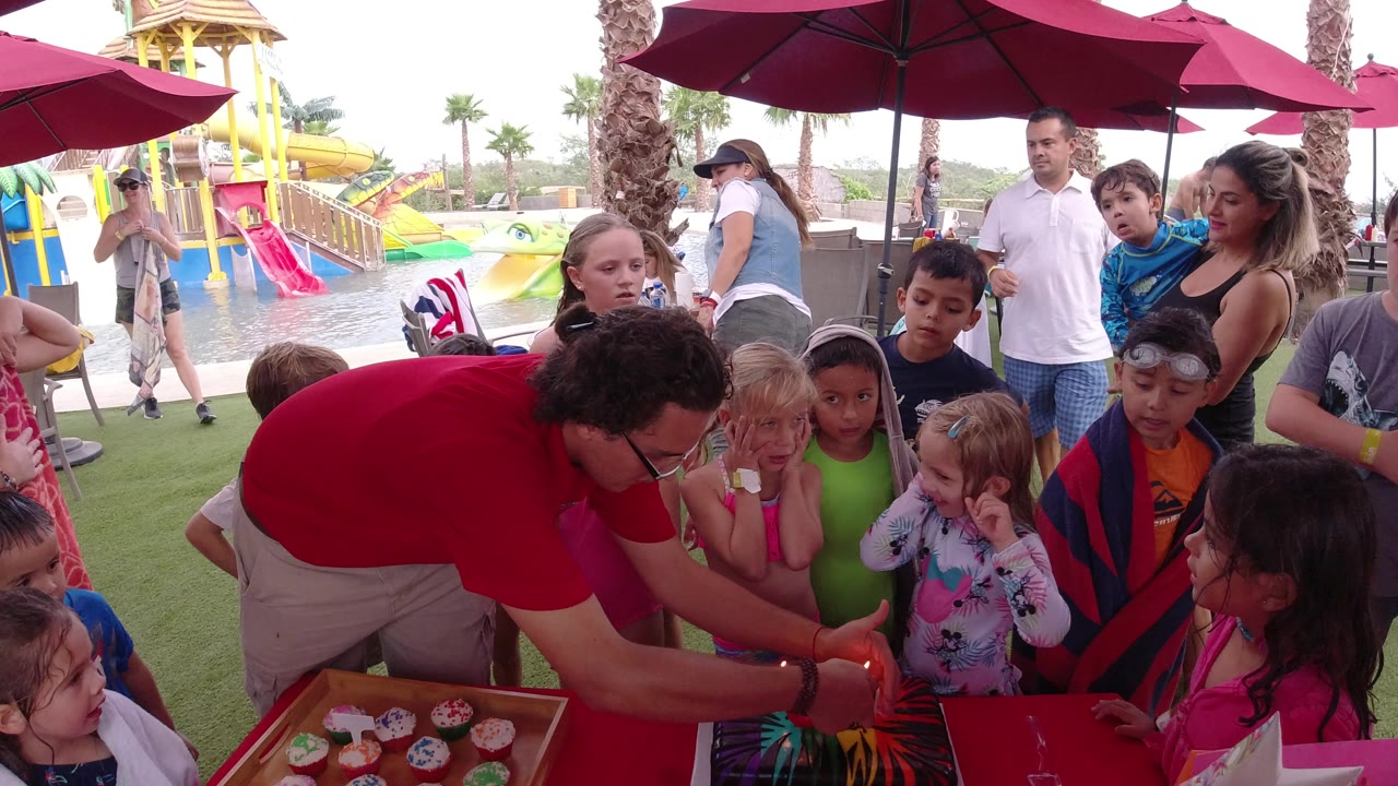 At 0:01, the man in red bends toward a young girl at the party table while other children watch and colorful water slides sit behind them in Los Cabos.