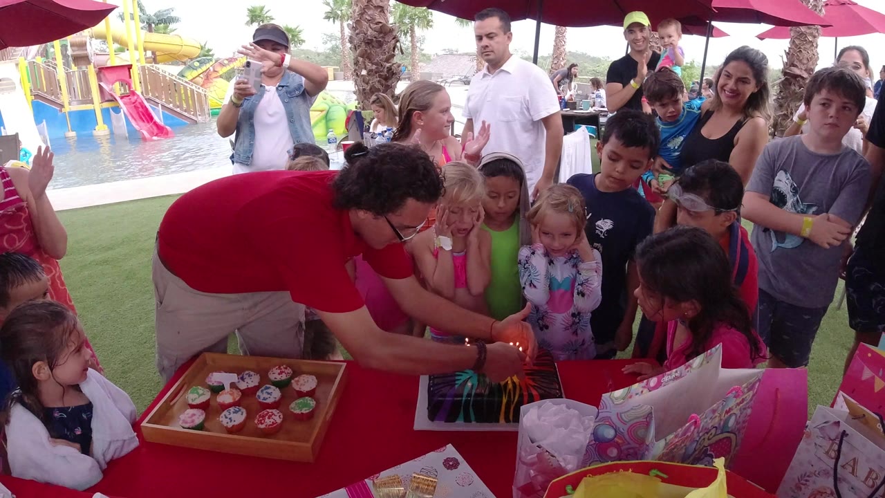 Children and adults gather around a birthday cake on an outdoor party table.
