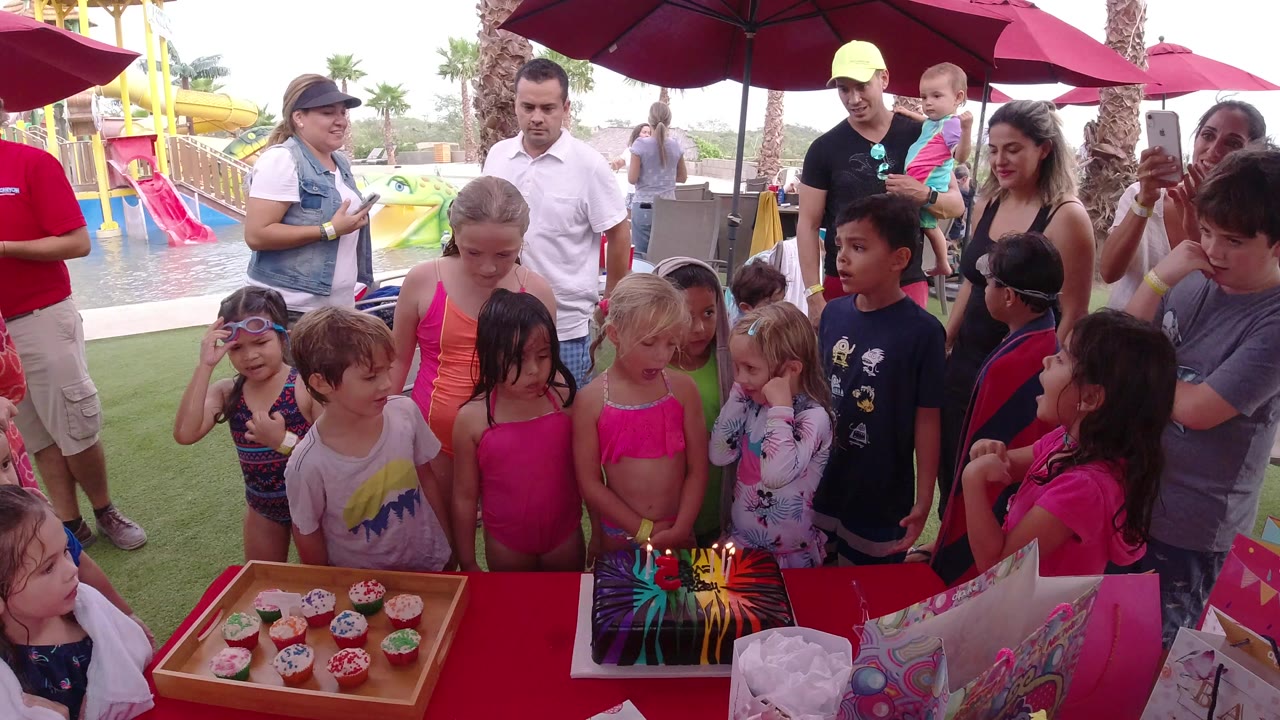 Children gather around a birthday cake and cupcakes at an outdoor party.