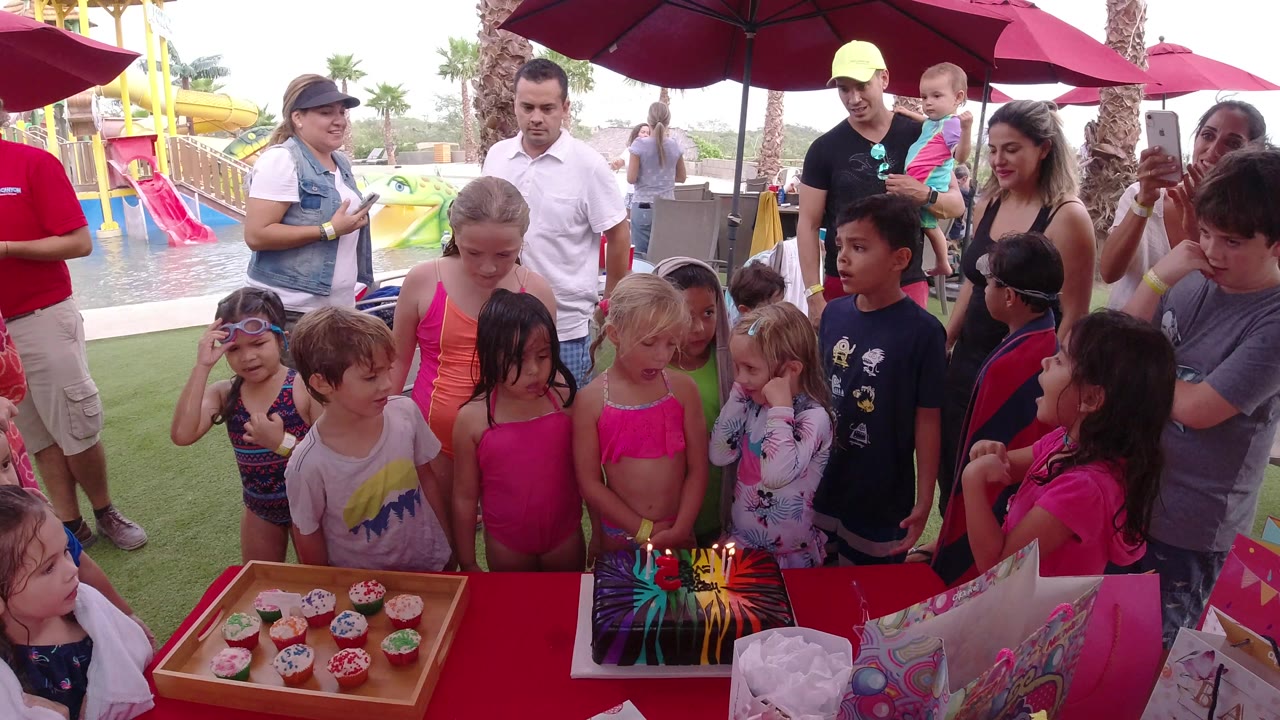 At 0:01 in Los Cabos, Mexico, the group remains around the red table, with the girl in a pink swimsuit near the cake and adults holding phones under red umbrellas.