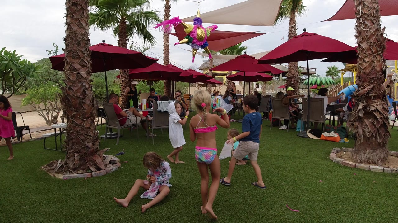 At the Los Cabos garden party, children shift positions on the lawn while adults remain seated under umbrellas and the bright pinata hangs above the center of the scene.