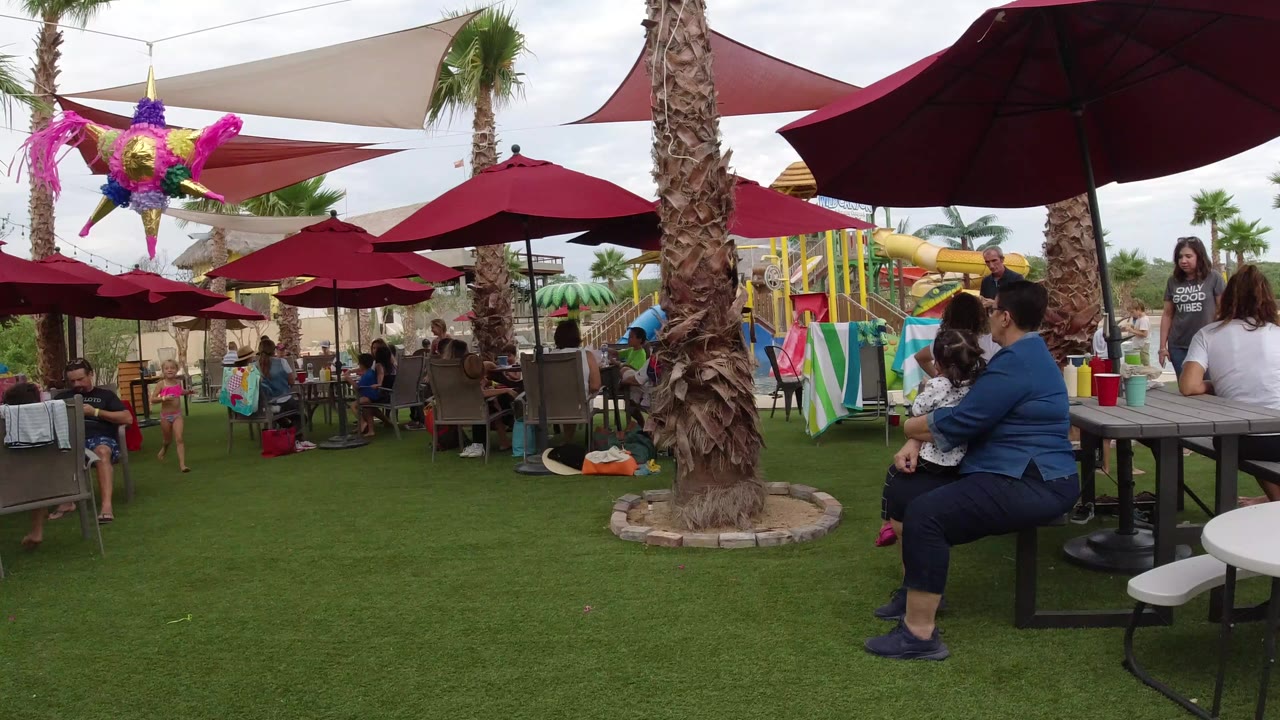 Resort patio with red umbrellas, palm trees, seated guests, and children on green turf.