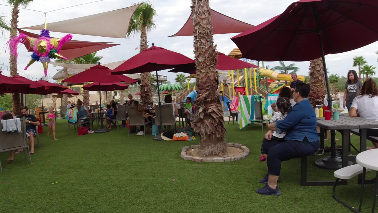 Los Cabos resort seating area with adults at tables under red umbrellas, children near the center, palm trunks, shade sails, and colorful play equipment behind them.