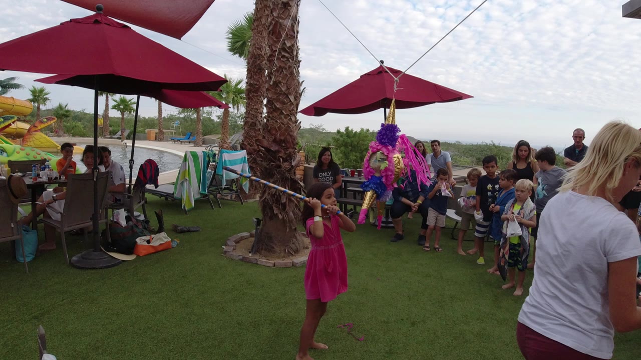 At a Los Cabos outdoor birthday party, a girl in a pink dress stands near a hanging pink and purple pinata while children and adults watch beneath red umbrellas.