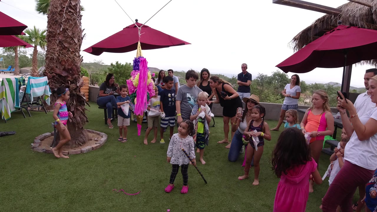 At a Los Cabos outdoor party, children and adults stand around a purple and pink pinata beneath burgundy umbrellas, with palm trunks and patio furniture in view.