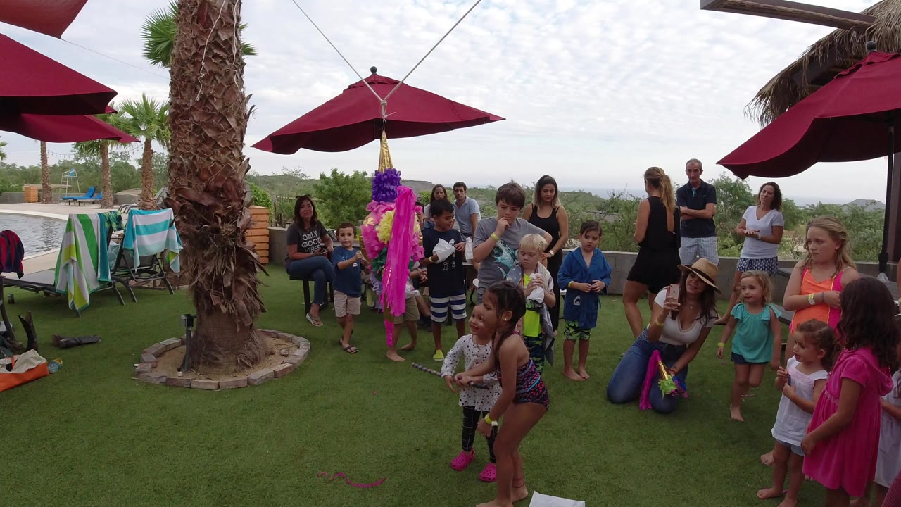 Children and adults gather around a colorful pinata on a patio lawn.