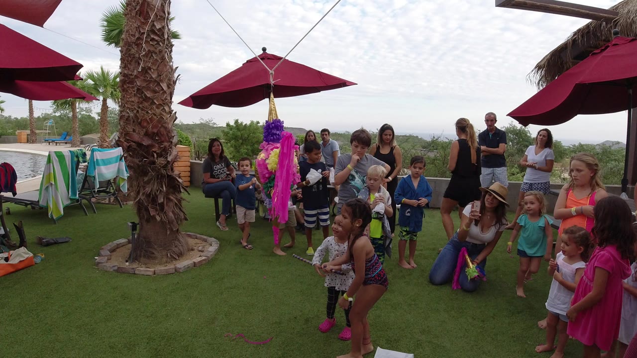 Adults and children in Los Cabos, Mexico, watch a pink and purple pinata hanging from lines between red umbrellas, with patio seating and greenery in the background.