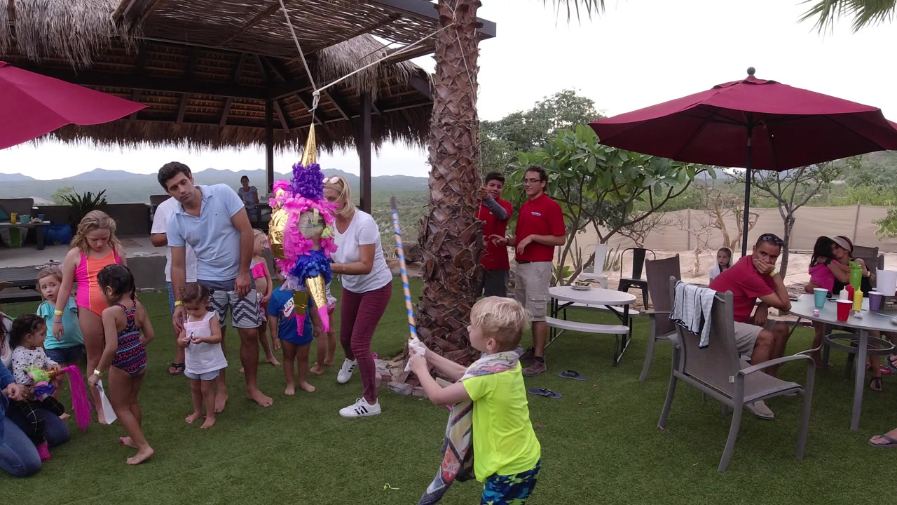 In Los Cabos, Mexico, children and adults stand on artificial grass around a bright hanging party figure under red umbrellas and a thatched roof.