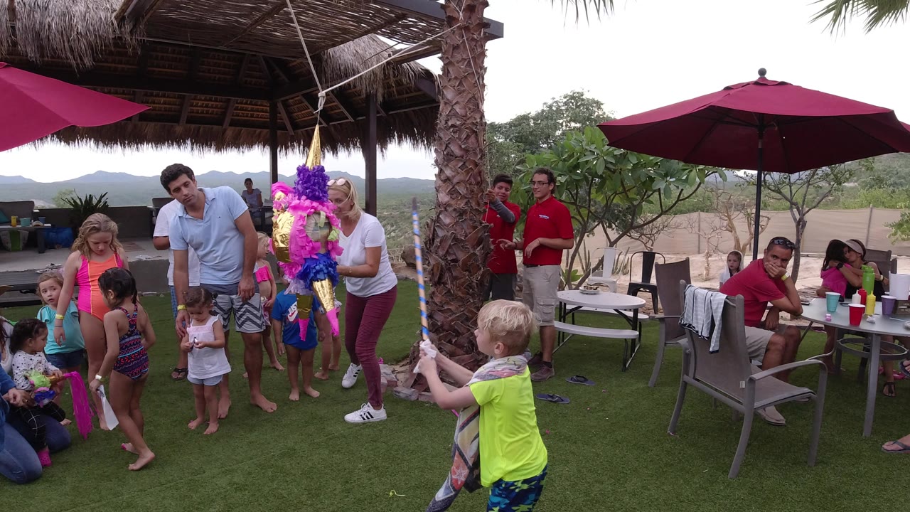 A child in a bright yellow shirt stands near the foreground with a stick pointed toward the colorful party figure as adults and children watch on the terrace in Los Cabos, Mexico.