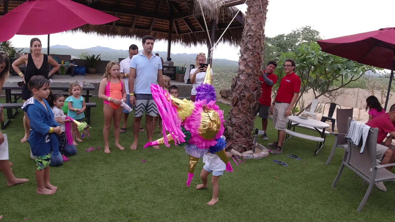 A little girl in a white top and shorts approaches the hanging pinata in Los Cabos as children and adults watch from the patio lawn.