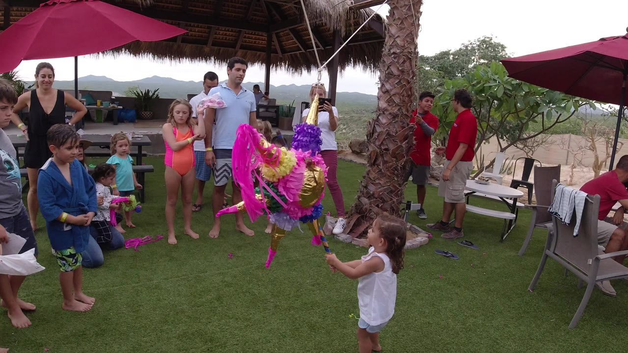 In Los Cabos, Mexico, children and adults stand on an outdoor turf patio as a young child faces a colorful piñata beneath red umbrellas and a thatched roof.