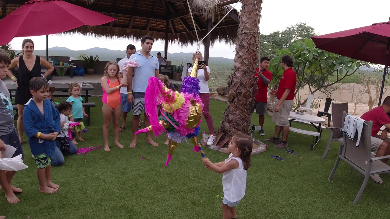In Los Cabos, Mexico, a young child in a white sleeveless top holds a stick toward the hanging pink and gold piñata as children and adults watch from the turf patio.